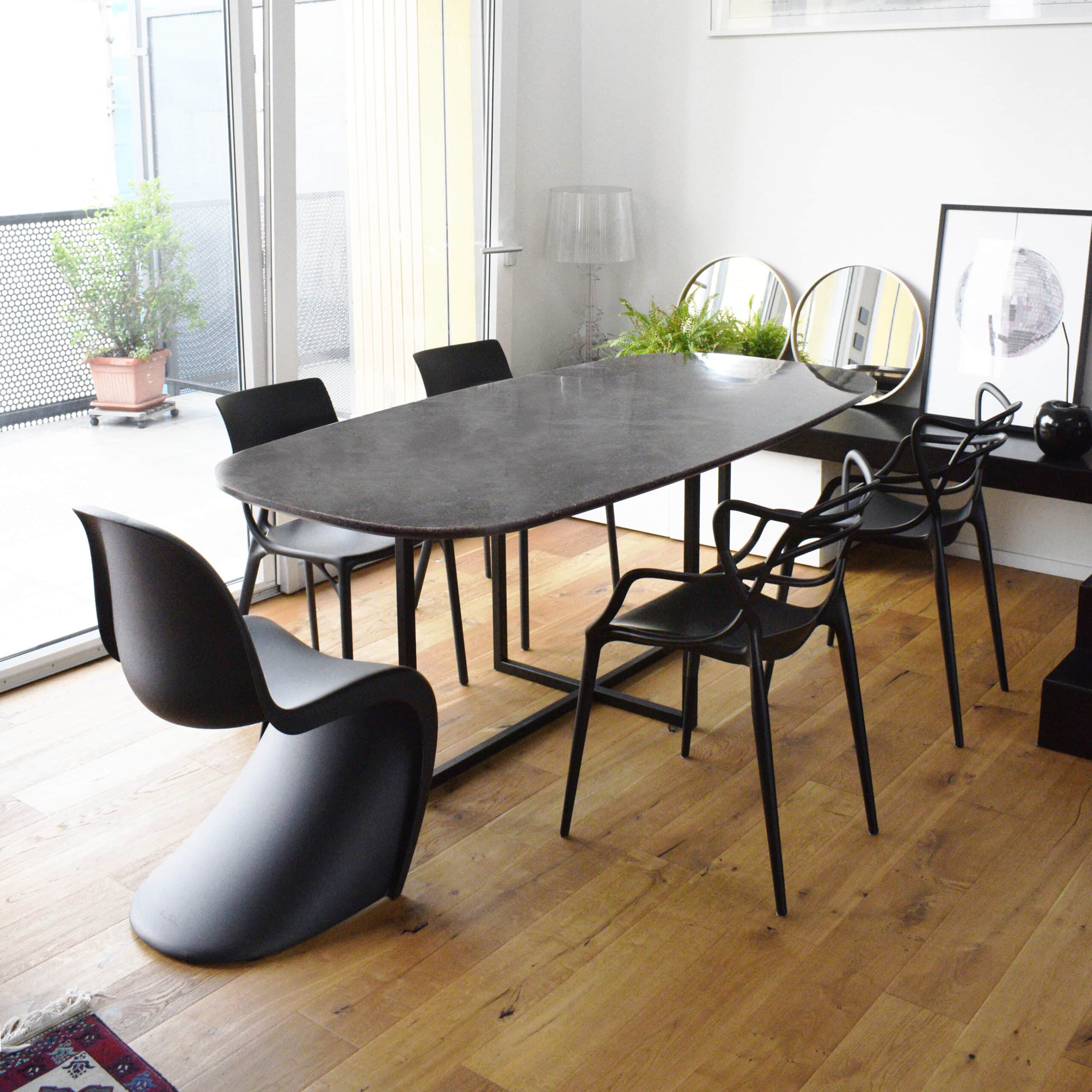 Un tavolo moderno con piano scuro in plastica 100% riciclata e riciclabile, circondato da sedie di design nero, in un ambiente luminoso con pavimento in legno. La stanza è arredata con elementi minimalisti, tra cui specchi decorativi e piante verdi.